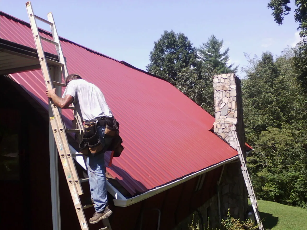 Roofer on a ladder beside a red metal roof and stone chimney — Soffit & Fascia Repair by Roofing Brothers in Finksburg